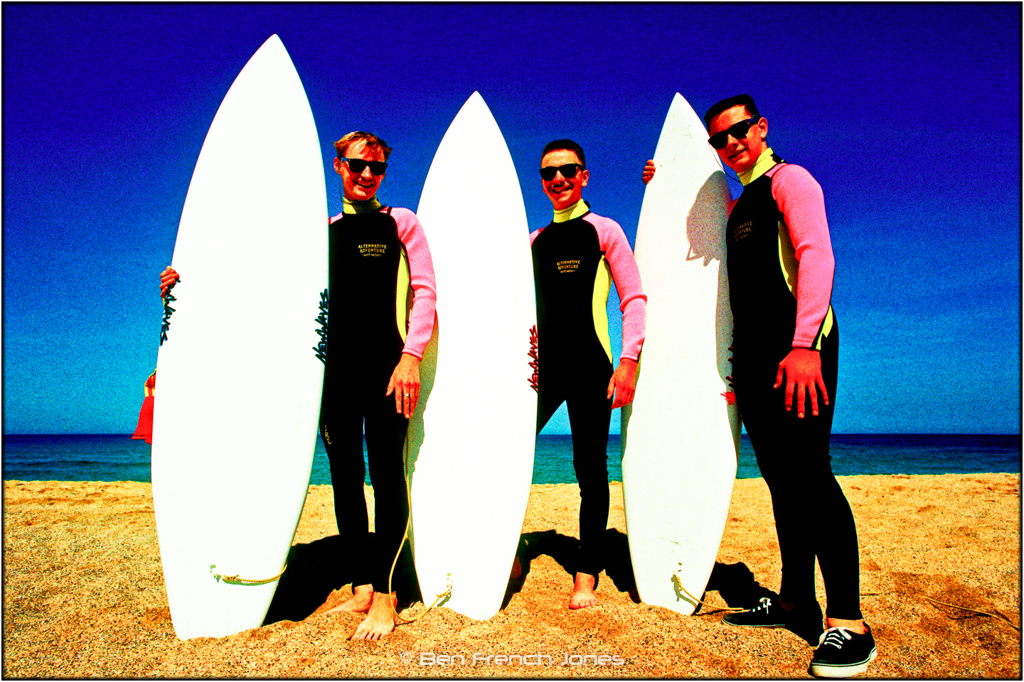 three guys in matching wetsuits standing with surfboards on a sunny beach